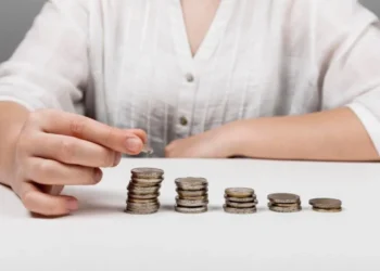 Mão de uma pessoa organizando várias pilhas de moedas em crescimento sobre uma mesa, simbolizando o conceito de equiparação salarial e igualdade de rendimentos.