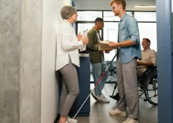 Um grupo de profissionais em um escritório inclusivo, com um homem usando bengala para mobilidade visual conversando com uma colega, enquanto outros funcionários, incluindo uma pessoa em cadeira de rodas, estão ao fundo.