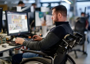 Homem em cadeira de rodas trabalhando em um computador em um escritório, com monitores e equipamentos modernos ao redor, representando a inclusão e acessibilidade no ambiente de trabalho.