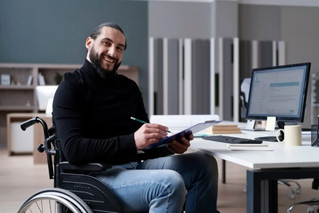 Homem em cadeira de rodas trabalhando em um escritório, sorrindo enquanto revisa documentos em uma prancheta, com um computador e materiais de trabalho na mesa.