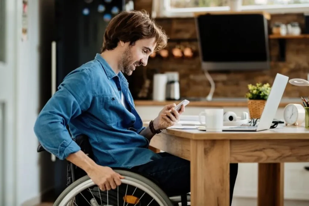 Homem em cadeira de rodas trabalhando de casa, usando um smartphone e um laptop em sua mesa, demonstrando acessibilidade e inclusão no ambiente de trabalho remoto.