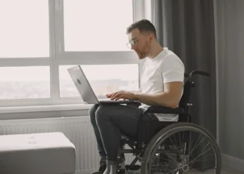 Homem em cadeira de rodas usando um laptop, sentado próximo a uma janela, em um ambiente doméstico iluminado.