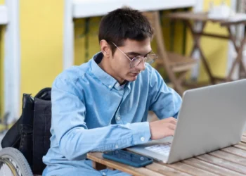 Jovem com óculos e camisa azul sentado em uma cadeira de rodas enquanto usa um laptop em uma mesa ao ar livre. A cena ocorre em um ambiente com fachada amarela e móveis de madeira.