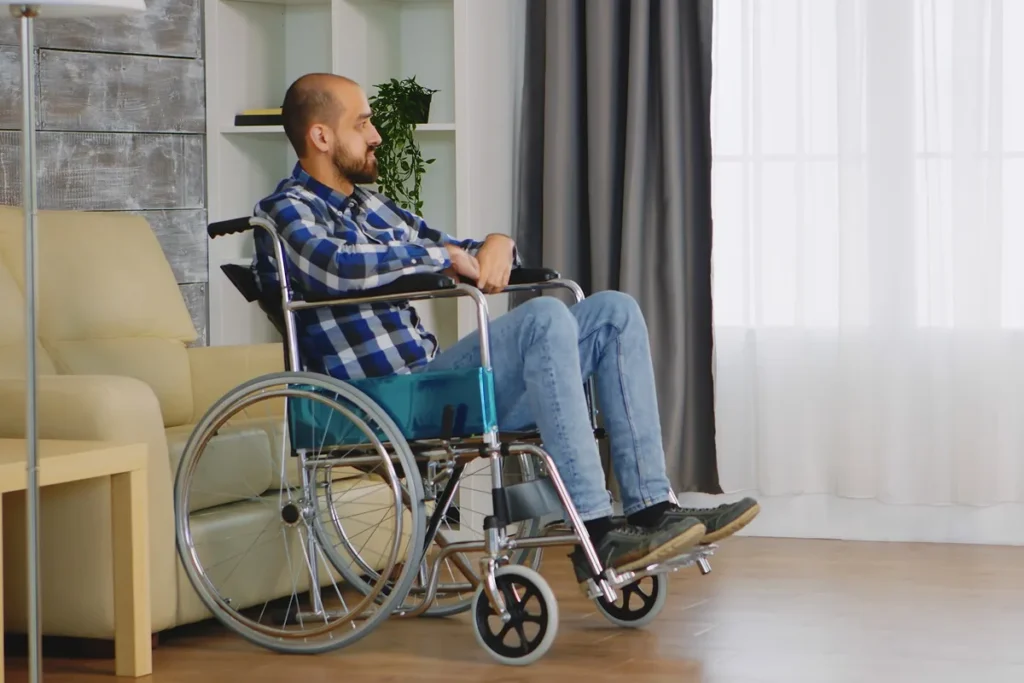 Homem sentado em uma cadeira de rodas, vestindo camisa xadrez azul e jeans, descansando em uma sala com sofá bege, cortinas claras e piso de madeira.