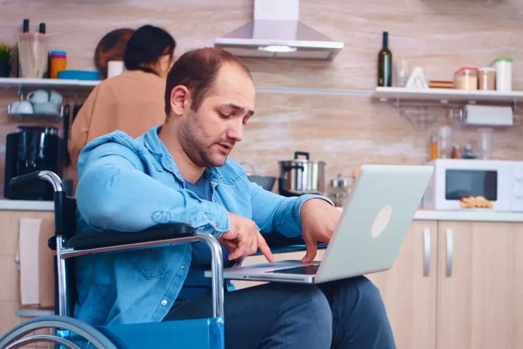 Homem em cadeira de rodas, vestido com camisa jeans, usando um laptop em uma cozinha moderna, enquanto uma mulher ao fundo realiza tarefas domésticas.