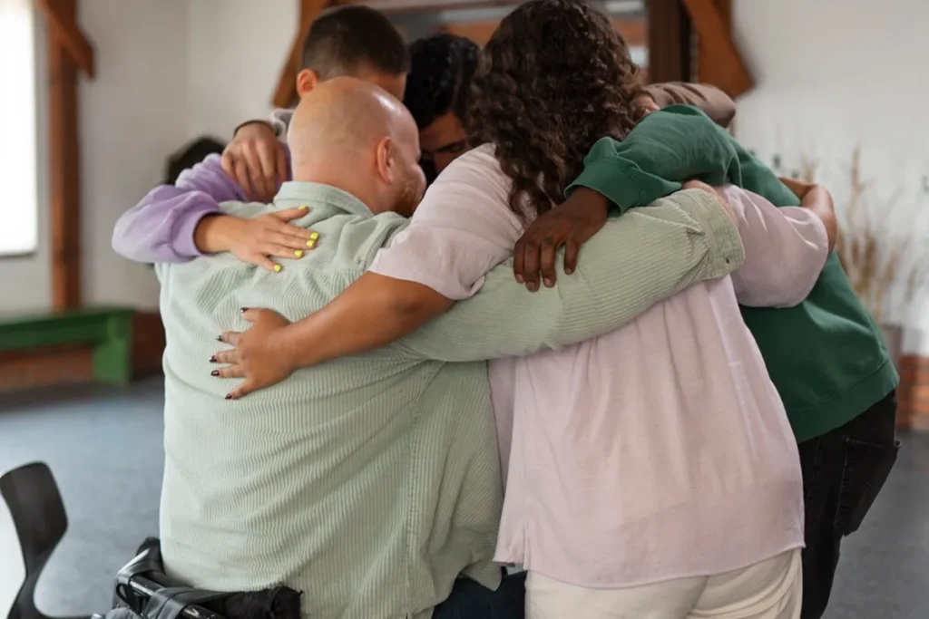 Grupo de pessoas de diversas etnias e idades em um abraço coletivo, incluindo uma pessoa em cadeira de rodas, promovendo união e inclusão em um ambiente comunitário.