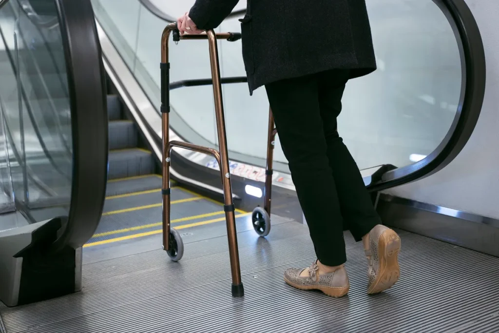 Uma pessoa com mobilidade reduzida utilizando um andador metálico ao subir uma escada rolante. A imagem mostra as adaptações e desafios enfrentados por pessoas com dificuldade de locomoção em ambientes urbanos.