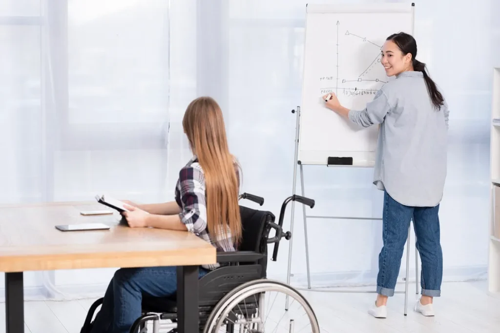 Mulher em cadeira de rodas assistindo a uma aula, enquanto uma instrutora escreve gráficos em um quadro branco. A cena destaca inclusão em ambientes educacionais.