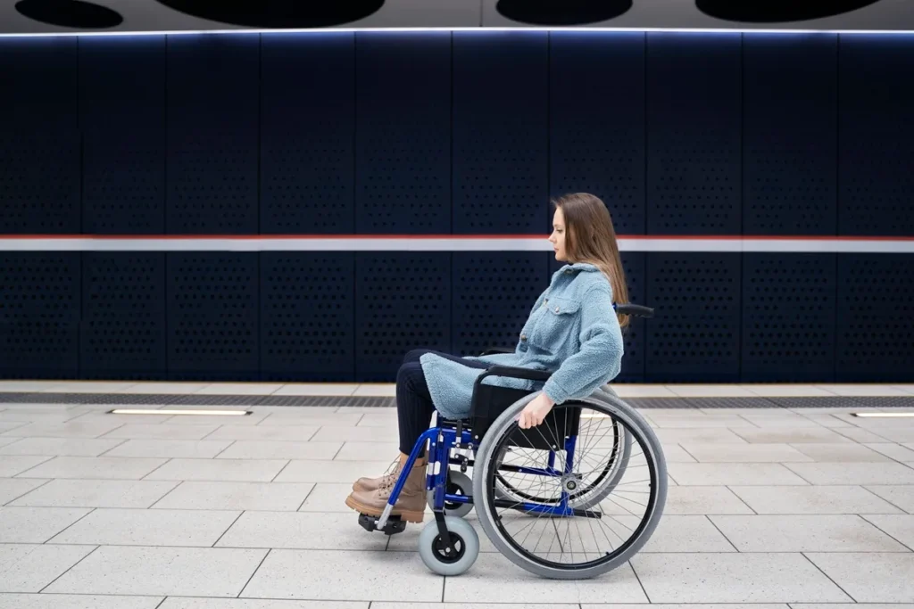 Mulher jovem, usando casaco azul e botas marrons, sentada em uma cadeira de rodas em uma estação de transporte público com design moderno e linhas minimalistas.