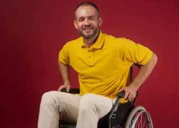 Homem usando camisa amarela e sentado em uma cadeira de rodas, sorrindo, com fundo vermelho.