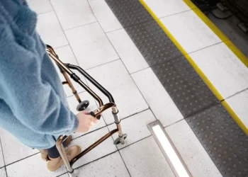 Uma pessoa usando um andador em uma plataforma de transporte público, próxima a uma faixa de segurança amarela e área de alerta tátil para deficientes visuais. A imagem destaca elementos de acessibilidade e segurança no ambiente urbano.