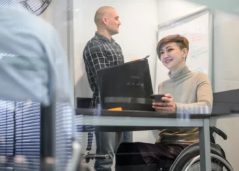 Mulher em cadeira de rodas trabalhando em equipe, usando laptop e segurando um copo, enquanto um colega faz anotações em um quadro branco em uma sala de reunião moderna.