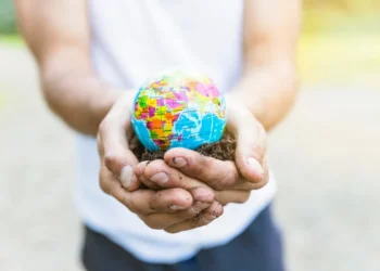 Mãos de uma pessoa segurando uma pequena réplica do globo terrestre sobre terra, simbolizando responsabilidade social e sustentabilidade.