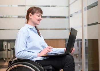 Mulher em cadeira de rodas, usando camisa social azul, trabalhando em um laptop em um escritório moderno com divisórias de vidro.