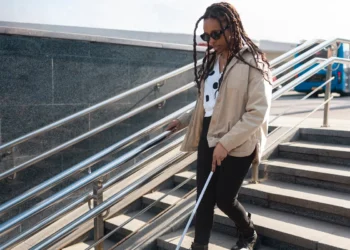 Uma mulher com deficiência visual, usando óculos escuros e uma bengala branca, descendo escadas em um ambiente urbano. A imagem destaca a importância de acessibilidade e infraestrutura adequada para pessoas com deficiência.