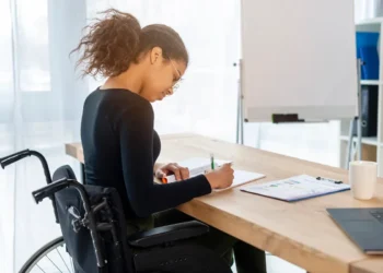Mulher em cadeira de rodas escrevendo em caderno sobre uma mesa de madeira, com gráficos e materiais de estudo ao lado, em um ambiente iluminado e profissional.