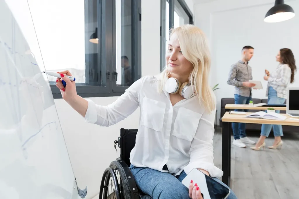 Mulher utilizando cadeira de rodas, escrevendo em um quadro branco em um ambiente de trabalho moderno, com colegas interagindo ao fundo.