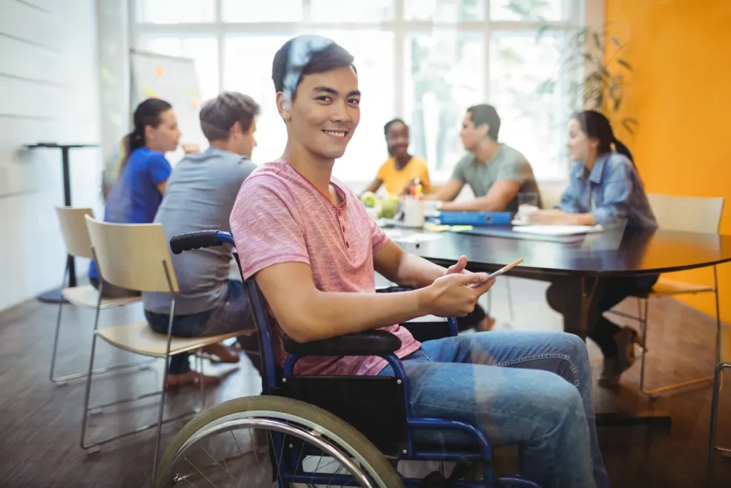 Jovem em cadeira de rodas sorrindo e segurando um tablet durante uma reunião de equipe em um ambiente de escritório moderno.