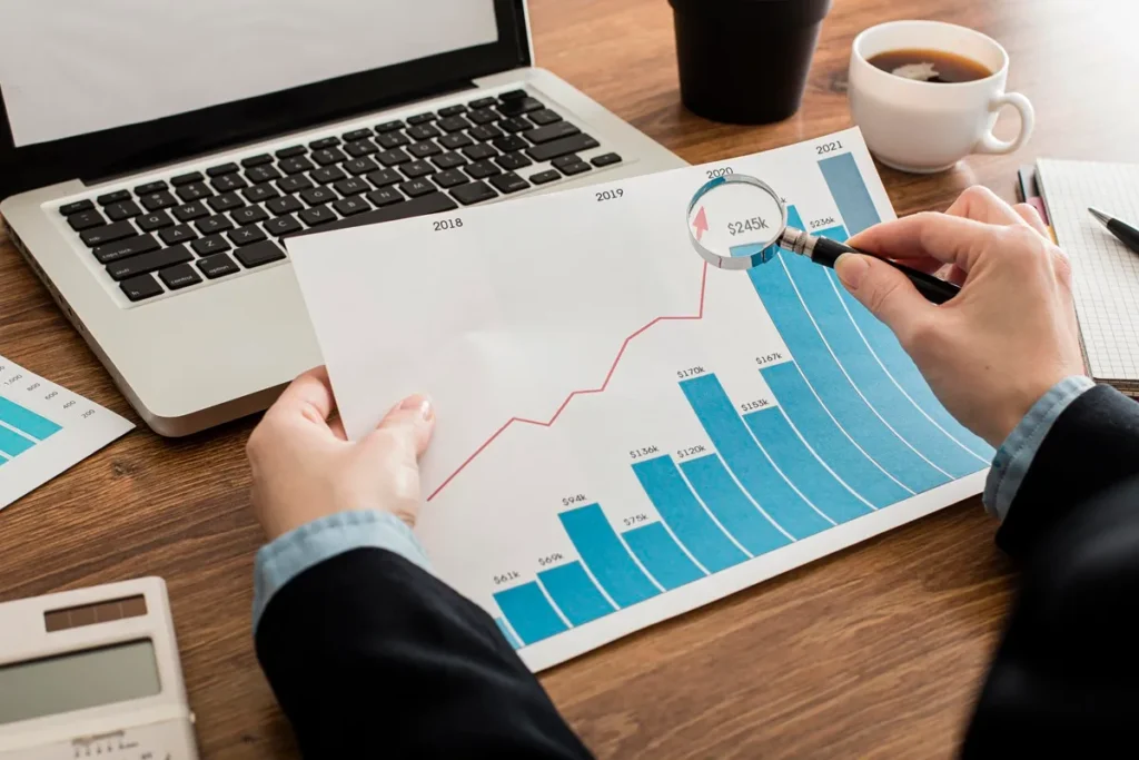 Pessoa analisando gráfico de crescimento financeiro com lupa, ao lado de um laptop, calculadora e xícara de café em uma mesa de madeira.