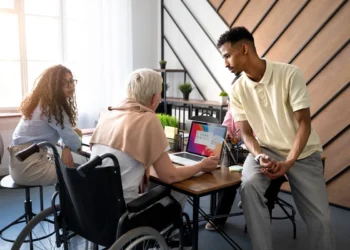 Equipe de trabalho diversificada reunida em um ambiente corporativo, discutindo estratégias de produtividade e redução do absenteísmo. Uma colaboradora em cadeira de rodas interage com colegas, destacando a importância da inclusão e do engajamento no ambiente profissional.
