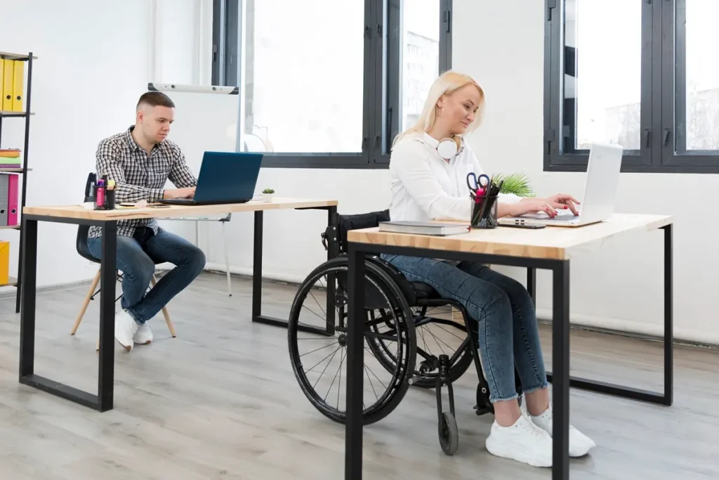 Mulher em cadeira de rodas trabalhando em um escritório moderno e acessível, ao lado de um colega concentrado em seu laptop. A imagem destaca a importância da inclusão no ambiente corporativo para reduzir o absenteísmo e aumentar a produtividade.