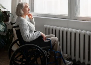 Idosa com cabelos grisalhos sentada em cadeira de rodas próxima a uma janela, olhando pensativa para fora.