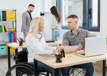 Mulher em cadeira de rodas trabalhando em equipe com colega em um escritório moderno e inclusivo, enquanto outros profissionais interagem ao fundo.