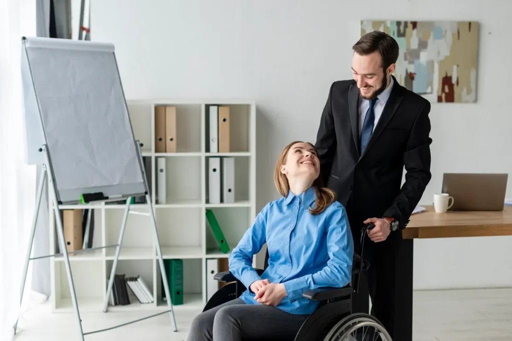 Mulher em cadeira de rodas e colega de trabalho de terno preto interagem com sorriso em um escritório moderno e acessível.