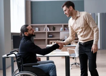 Homem em cadeira de rodas e colega de trabalho apertando as mãos em um escritório moderno, simbolizando inclusão e acessibilidade profissional.