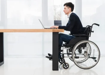 Homem cadeirante trabalhando em um laptop sobre uma mesa em um ambiente corporativo moderno e acessível.