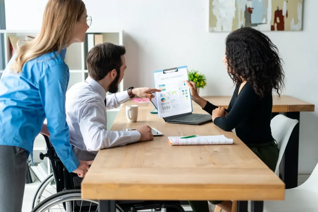Equipe corporativa em reunião, incluindo um profissional cadeirante, analisando gráficos e estratégias de mercado em um ambiente de trabalho moderno.