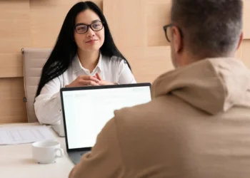 Mulher de óculos e camisa branca conduz entrevista com candidato em escritório, ambos sentados frente a frente com notebook sobre a mesa.