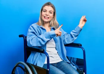 Mulher jovem e sorridente, com cabelos loiros e pele clara, sentada em uma cadeira de rodas azul. Ela veste uma camisa azul sobre uma blusa branca e calça jeans. Aponta para o lado com os dois dedos, como se estivesse explicando algo ou apresentando um conceito. O fundo é de um tom azul vibrante, criando um visual harmônico e moderno.