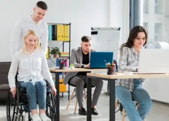 Equipe diversa trabalhando em um escritório moderno, com uma mulher em cadeira de rodas sendo auxiliada por um colega.