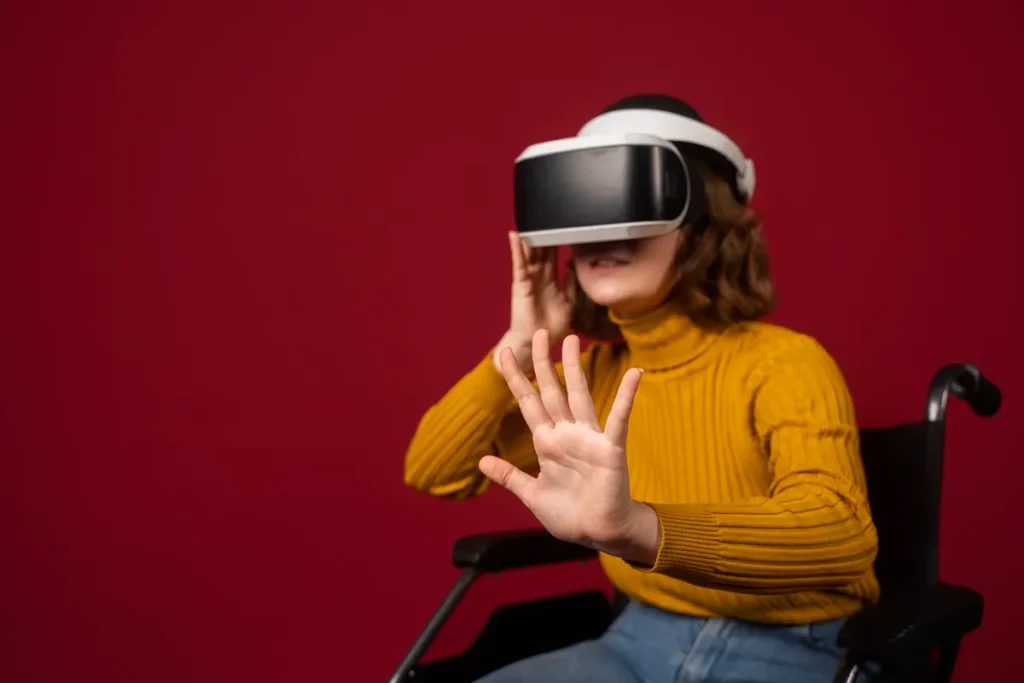 Mulher sentada em cadeira de rodas usando óculos de realidade virtual, com um suéter amarelo e fundo vermelho, interagindo com o ambiente digital ao estender a mão para a frente.