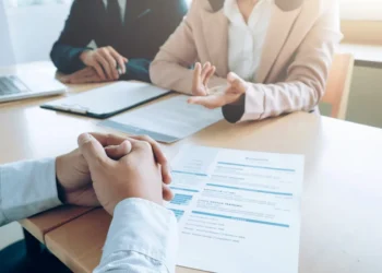 Candidato participando de entrevista de emprego com currículo sobre a mesa e recrutadores ao fundo