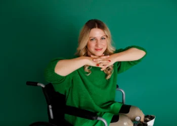 Mulher com deficiência física, usando cadeira de rodas e prótese nas pernas, sorrindo com as mãos apoiadas sobre o rosto, vestida com roupa verde e posando em frente a um fundo da mesma cor.