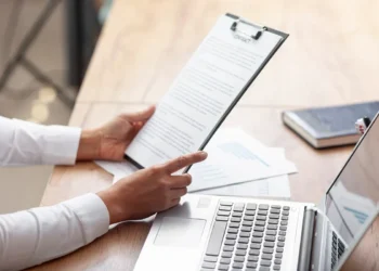Pessoa segurando carta de apresentação em prancheta, com laptop e documentos na mesa