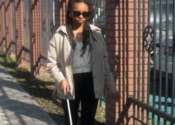 Imagem de uma mulher com óculos de sol, usando um bastão de mobilidade enquanto caminha em uma rua ao ar livre, com cercado de tijolos e carros ao fundo