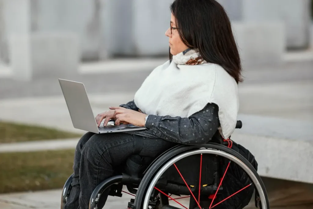 Imagem de uma mulher em cadeira de rodas, vestindo uma blusa escura e um xale branco, usando um laptop em um ambiente ao ar livre, com foco na acessibilidade e inclusão.