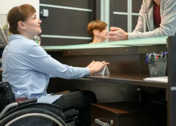 Mulher em cadeira de rodas sendo atendida em um balcão de atendimento, interagindo com um atendente.