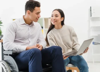 Homem em cadeira de rodas e mulher sorrindo enquanto olham para um laptop em um ambiente de trabalho moderno.